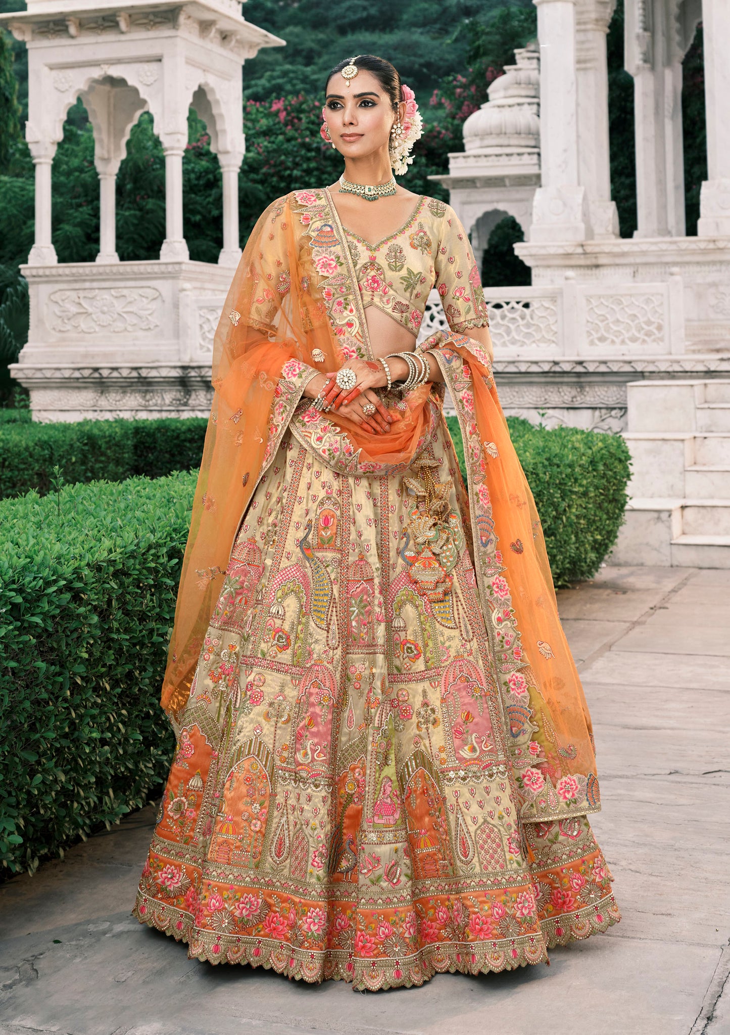 Woman in traditional embroidered outfit with orange dupatta standing in front of a white architectural structure.